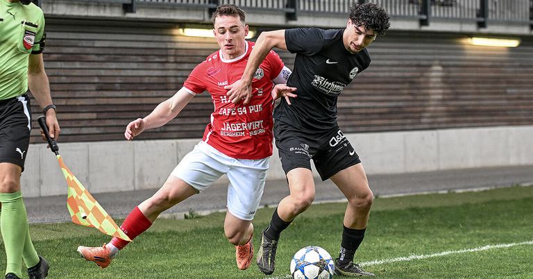 Two male soccer players are playing on a grass field. One is wearing a red jersey, and the other is wearing a black jersey. They are both trying to get the ball.