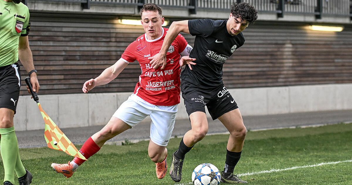 Two male soccer players are playing on a grass field. One is wearing a red jersey, and the other is wearing a black jersey. They are both trying to get the ball.