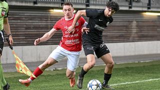 Two male soccer players are playing on a grass field. One is wearing a red jersey, and the other is wearing a black jersey. They are both trying to get the ball.