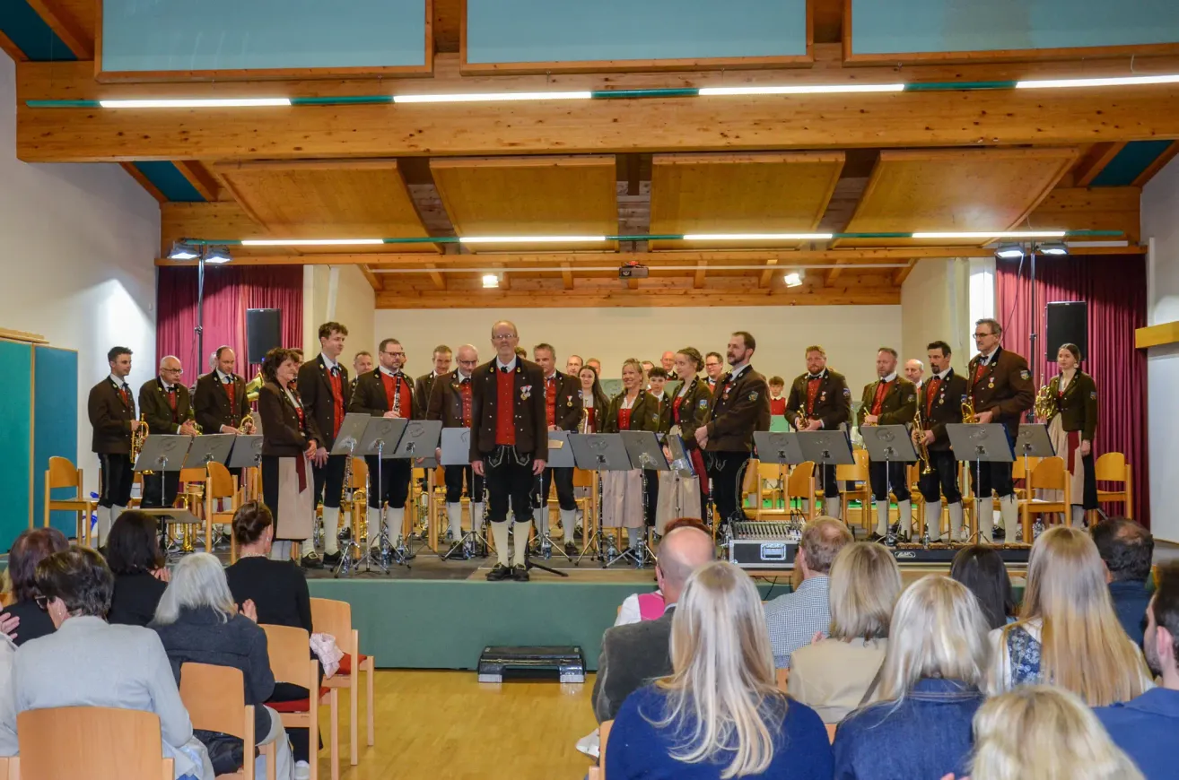 A brass band is performing on stage, with musicians in traditional attire. The audience is seated in front of them, watching the performance. The stage has music stands and equipment. The room has wooden beams and curtains.