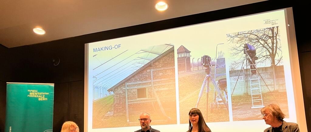 Eine Gruppe von Menschen ist drinnen und sitzt vor einer Projektionsleinwand. Die Leinwand zeigt ein Gebäude und die Worte 'MAKING-OF'. Die Menschen beobachten die Leinwand.