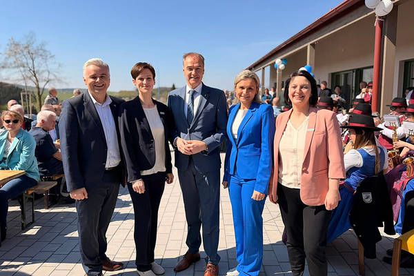 Fünf Erwachsene stehen zusammen und lächeln für ein Foto, gekleidet in formeller Kleidung. Sie sind draußen mit einem Gebäude im Hintergrund und einer großen Menschenmenge hinter ihnen.