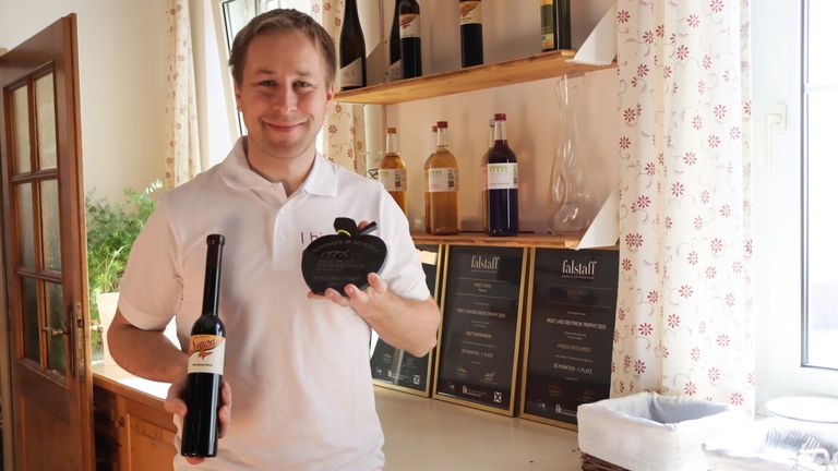 A man in a white polo shirt holds a bottle of wine and a trophy. Behind him, shelves with various bottles and a vase are displayed. Awards and certificates are mounted on the wall.