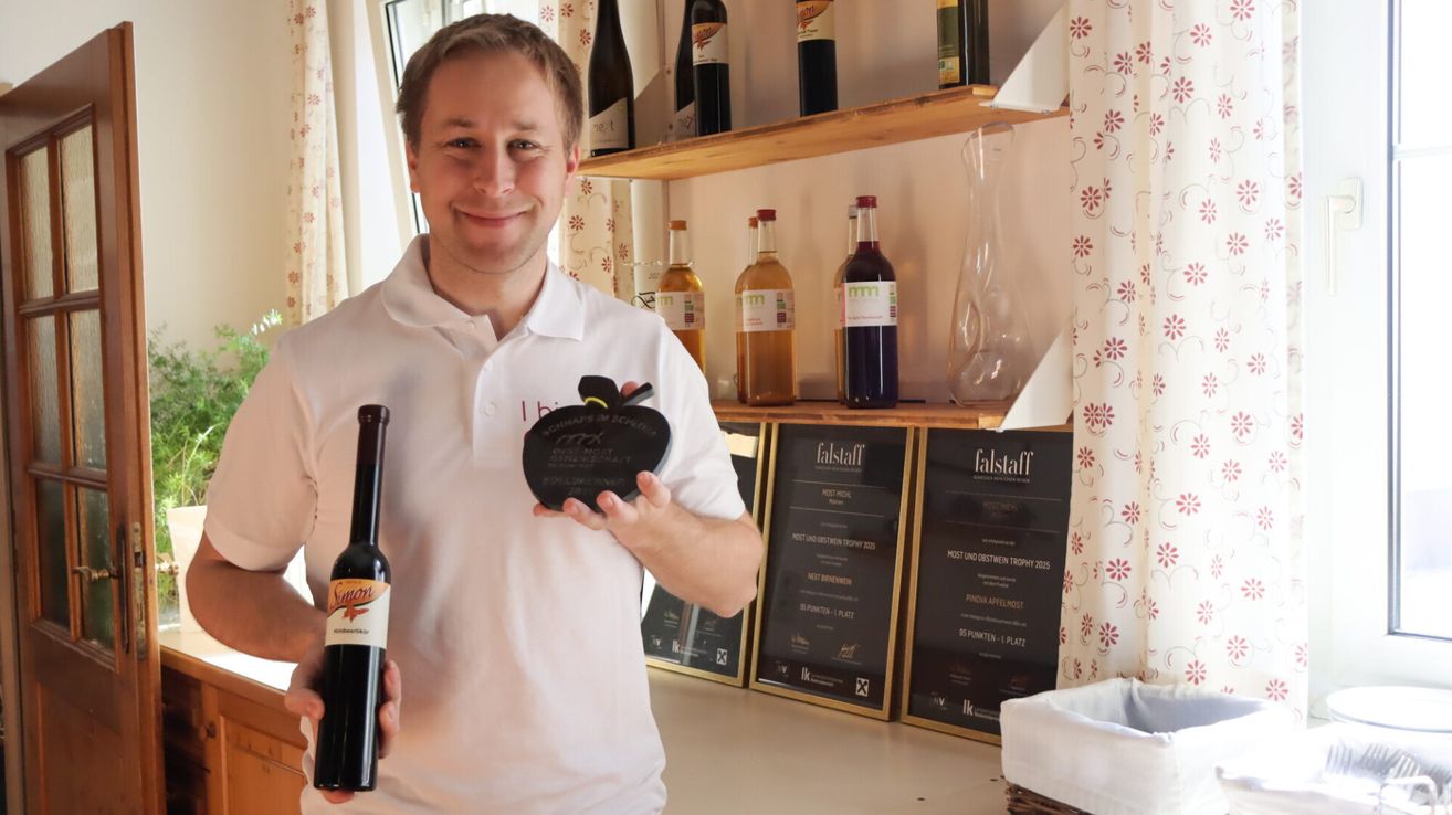 A man in a white polo shirt holds a bottle of wine and a trophy. Behind him, shelves with various bottles and a vase are displayed. Awards and certificates are mounted on the wall.