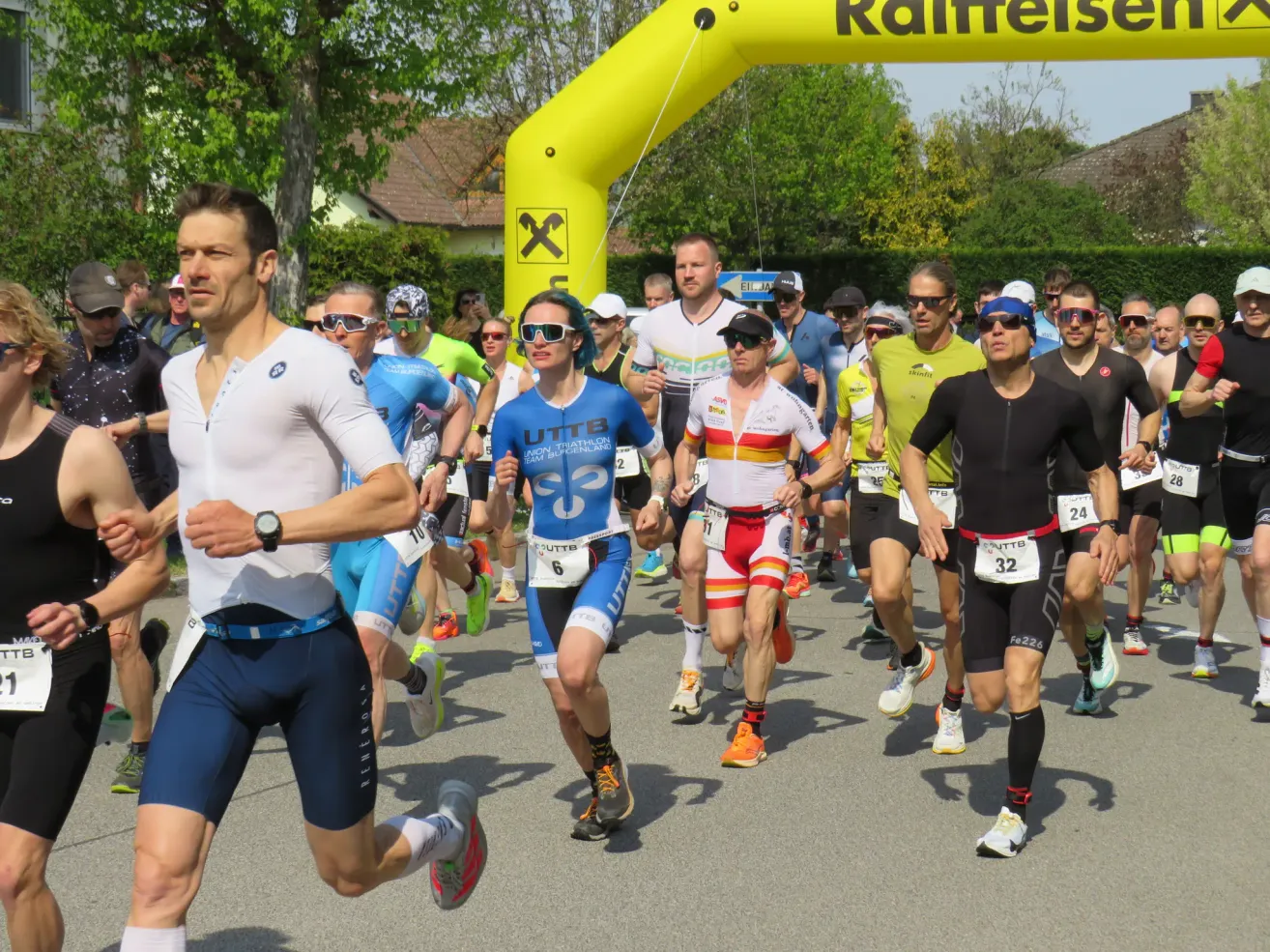 Eine Gruppe von Athleten läuft bei einem Marathonrennen und passiert dabei einen gelben Bogen mit der Aufschrift Raiffeisen. Sie tragen verschiedene Farben und einige von ihnen haben Sonnenbrillen auf. Der Wettkampf findet auf einer Straße statt.