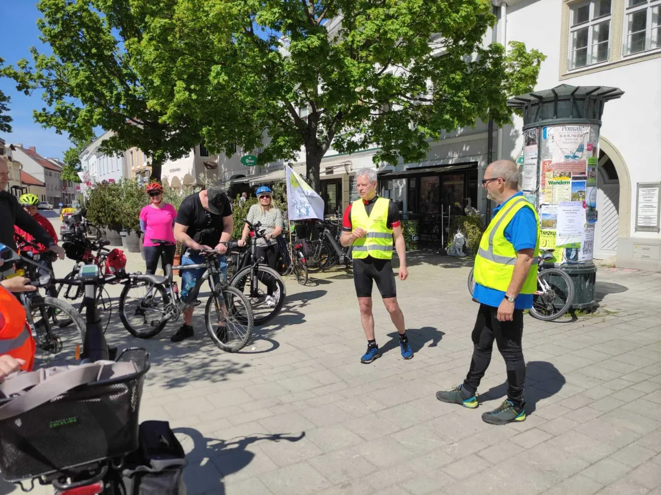 A group of cyclists are gathered in a town square, with one man speaking while another fixes his bike. Trees and buildings are in the background.