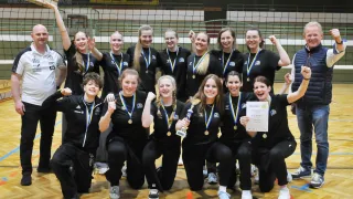 Eine Gruppe von Volleyballspielerinnen in schwarzen Uniformen mit Medaillen posiert für ein Foto auf einem Spielfeld. Sie halten eine Trophäe und ein Zertifikat in den Händen. Das Spielfeld hat ein Netz in der Mitte und Markierungen auf dem Boden.