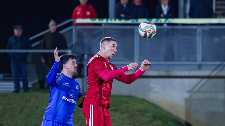 A soccer player in red is airborne, attempting to head the ball as another player in blue looks on. Spectators are watching from the bleachers.