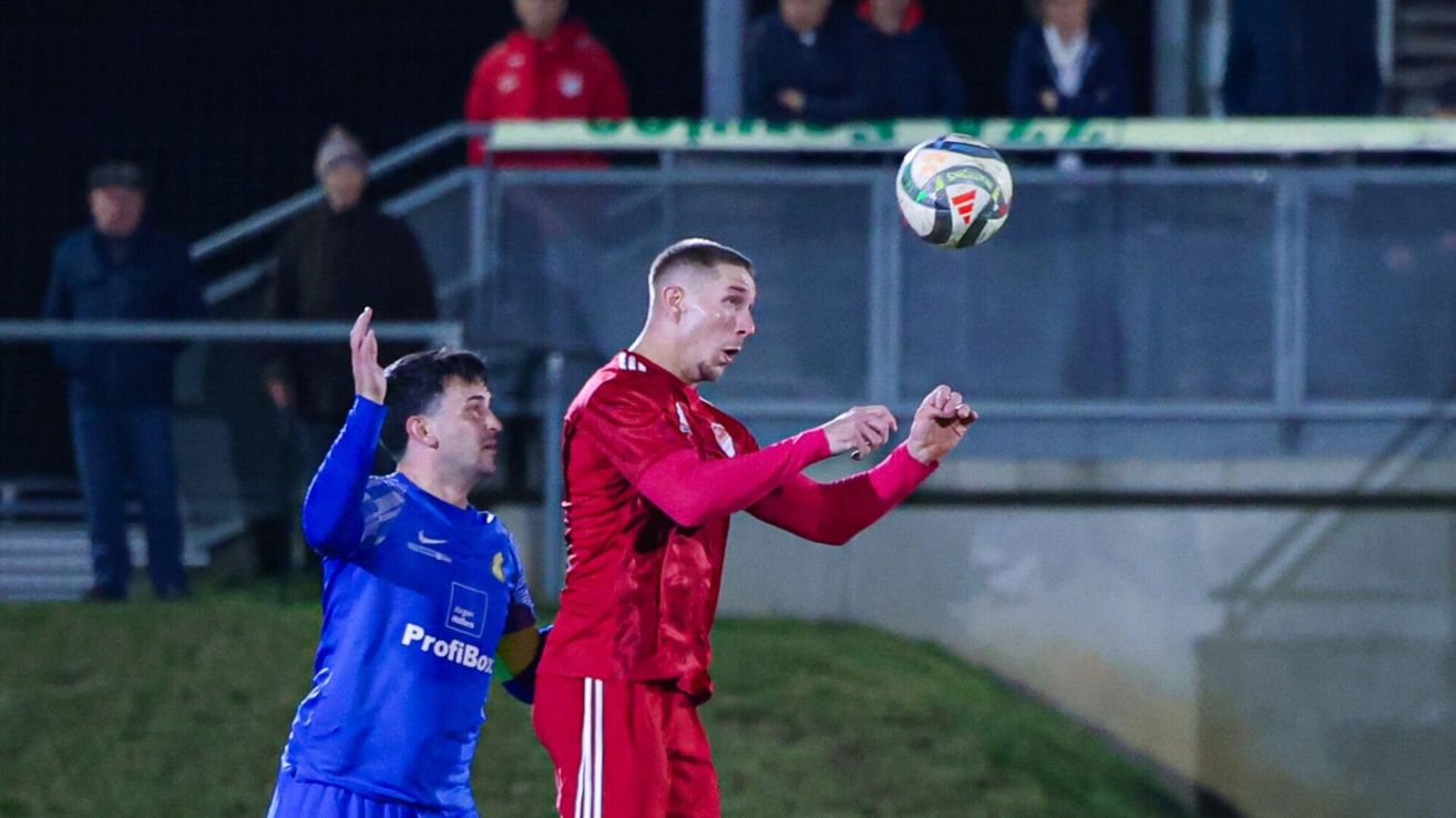 A soccer player in red is airborne, attempting to head the ball as another player in blue looks on. Spectators are watching from the bleachers.