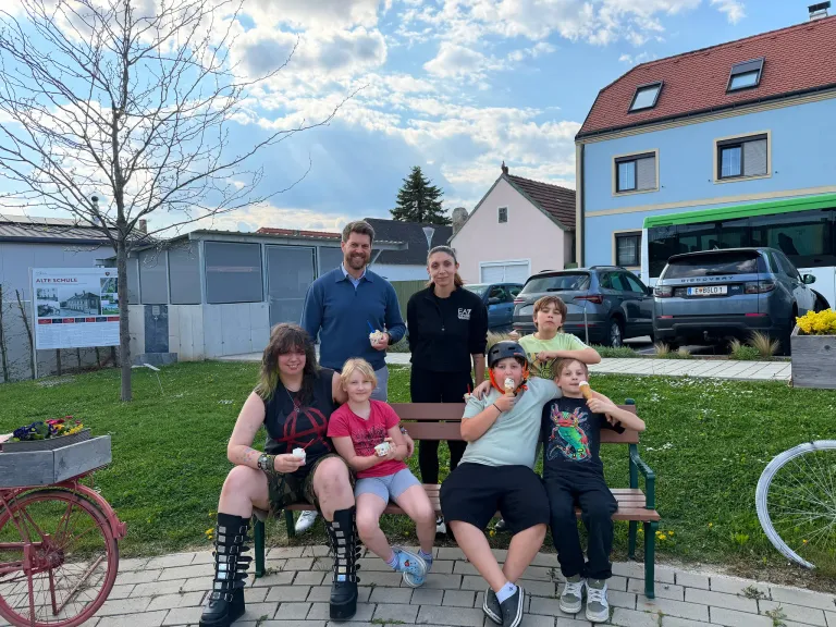 Eine Familie mit sieben Personen sitzt auf einer Parkbank. Sie essen alle Eis. Hinter ihnen stehen Gebäude, Autos und ein Bus. Der Himmel ist bewölkt.