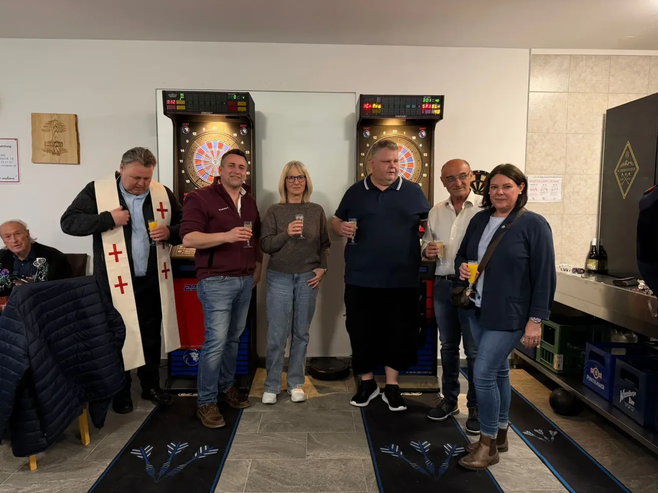 Eine Gruppe von Menschen steht vor Dartpfeilen und hält Getränke. Ein Mann trägt ein religiöses Gewand mit einem Kreuz, während ein anderer das Gewand eines Priesters trägt.