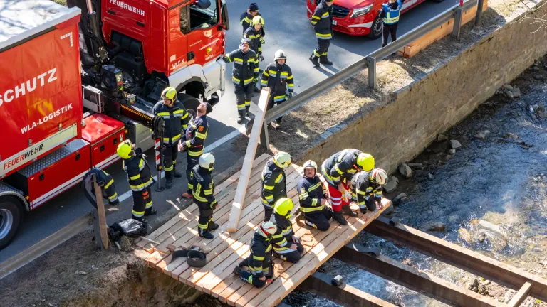 Firefighters in uniform work on a wooden bridge above a stream with a red fire truck in the background.