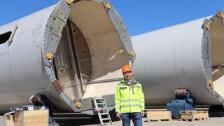 Ein Mann in einer reflektierenden Weste und einem Schutzhelm steht vor einer großen, offenen Metallstruktur, möglicherweise einer Windturbine, unter einem klaren blauen Himmel.