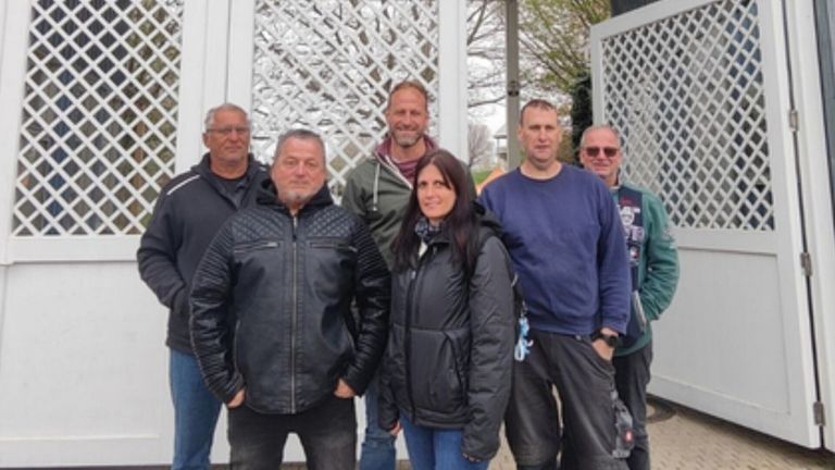 A group of people stands outside, smiling for a photo. A woman in a black jacket and two men in blue and green jackets are in the center.