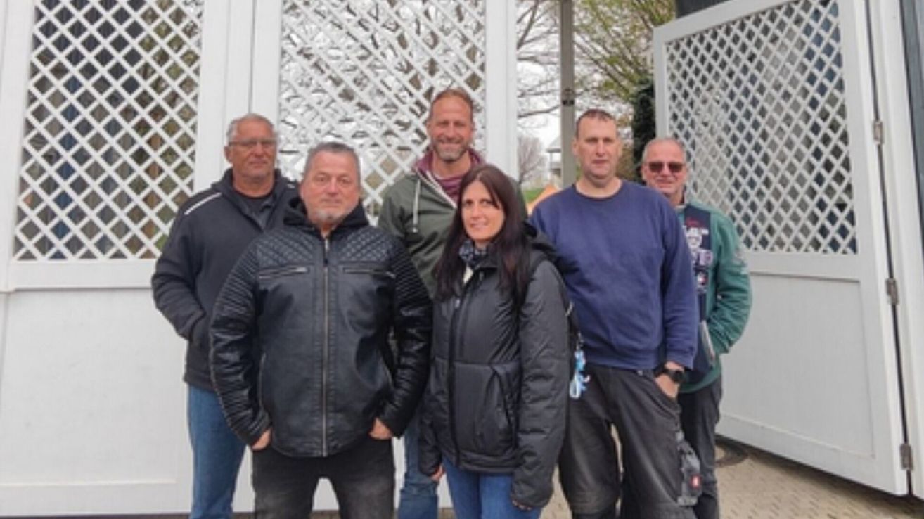A group of people stands outside, smiling for a photo. A woman in a black jacket and two men in blue and green jackets are in the center.