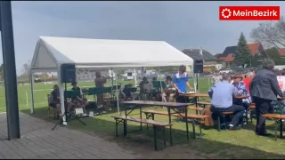 Eine Gruppe Musiker spielt unter einem Zelt mit Banner und Lautsprechern. Sie sitzen an Tischen mit Bänken. Ein Mann in einem blauen Shirt steht in der Nähe.