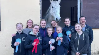 Eine Gruppe lächelnder junger Frauen und Männer steht vor einem weißen Pferd mit blauen und roten Bändern in der Hand. Das Pferd steht hinter ihnen in einem Stall.