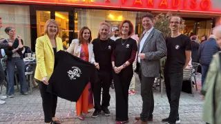 Fünf Personen stehen vor einem Neonschild mit der Aufschrift Cinema Paradis und halten ein schwarzes T-Shirt mit Text und Symbolen. Eine Frau in einem gelben Blazer hält ein schwarzes T-Shirt.