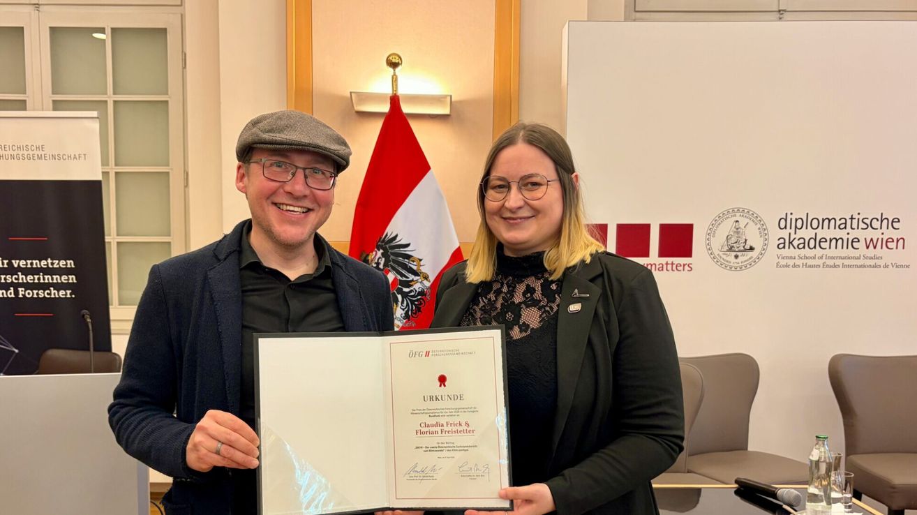 Ein Mann und eine Frau stehen lächelnd zusammen und halten ein Zertifikat. Der Mann trägt eine Mütze und eine Brille, die Frau eine Brille und ein schwarzes Spitzentop. Hinter ihnen ist eine Flagge und ein Schild mit roten Quadraten zu sehen.
