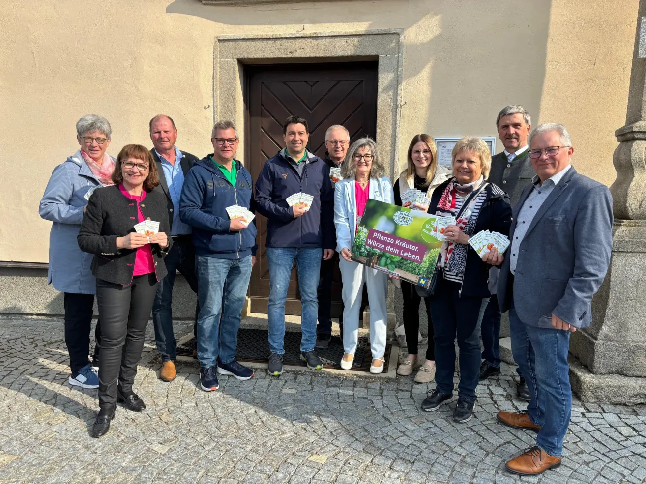 Group of adults standing in front of a door. They hold coupons and a sign that says 'Kurze Wege, kürzeres Leben.' They all smile and seem happy.