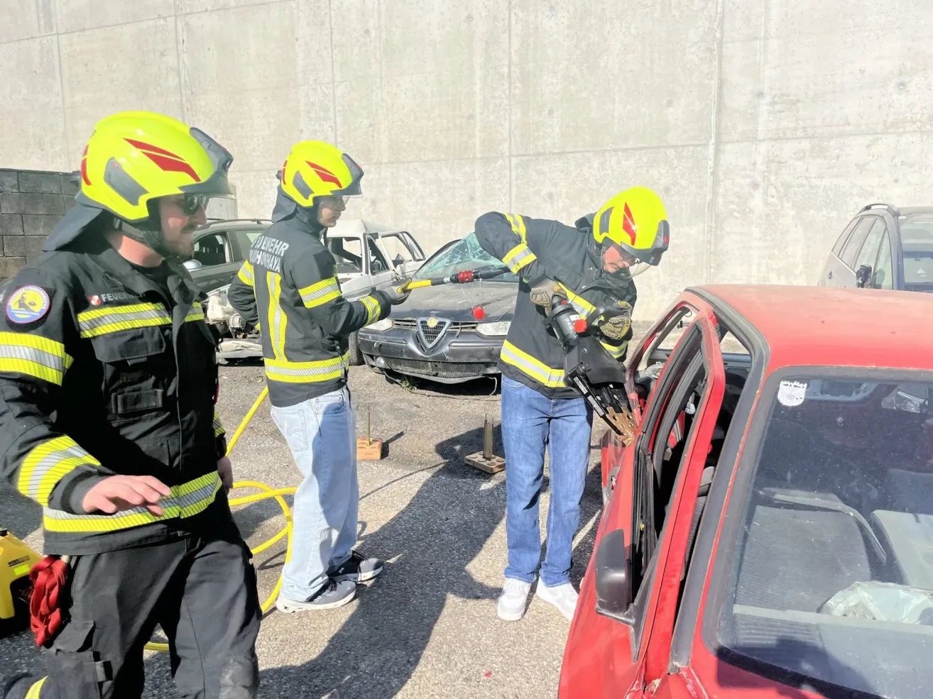 Three firefighters are outside, using tools on a red car with a damaged window. The car is parked next to a concrete wall, with other vehicles in the background.