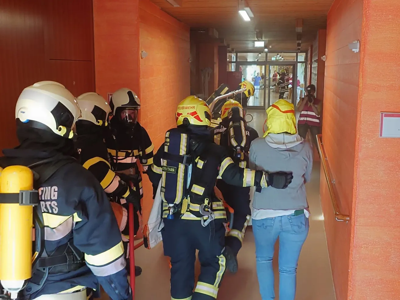 Firefighters in protective gear walk down a hallway with an adult and a child. The corridor is orange and well lit.