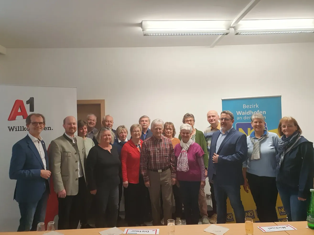 A group of people is standing in a conference room, posing for a photograph. A desk is in front of them with glasses and papers on it. There is a banner behind them.