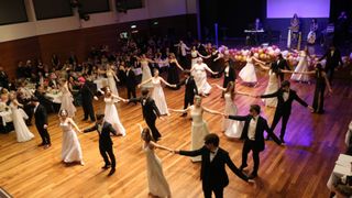 A large group of people in formal attire dancing in a circle on a stage. The audience is watching them.