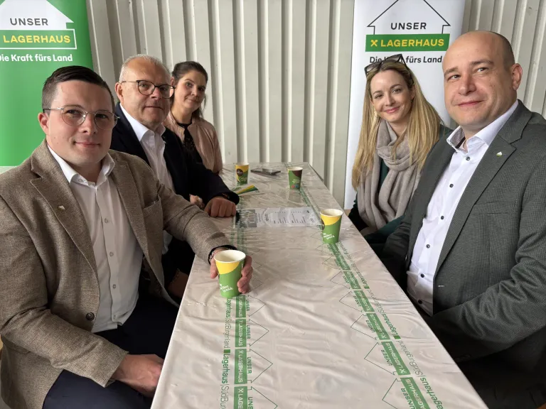 Fünf Personen sitzen mit grünen Bechern und einer weißen Tischdecke an einem Tisch und lächeln für ein Foto. Eine Frau hat eine Brille auf dem Kopf. Ein Banner hinter ihnen hat grünen Text.