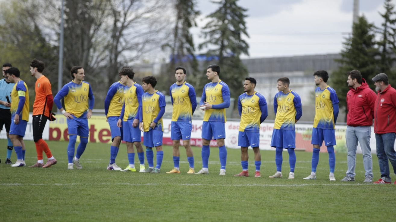 Sieben Fußballspieler stehen auf einem grünen Feld. Sie tragen gelbe und blaue Trikots. Ein Spieler hält einen Ball.