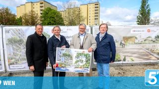 Four men stand at a construction site with a poster of a building design. They smile for a photo. Behind them, banners and buildings are visible.