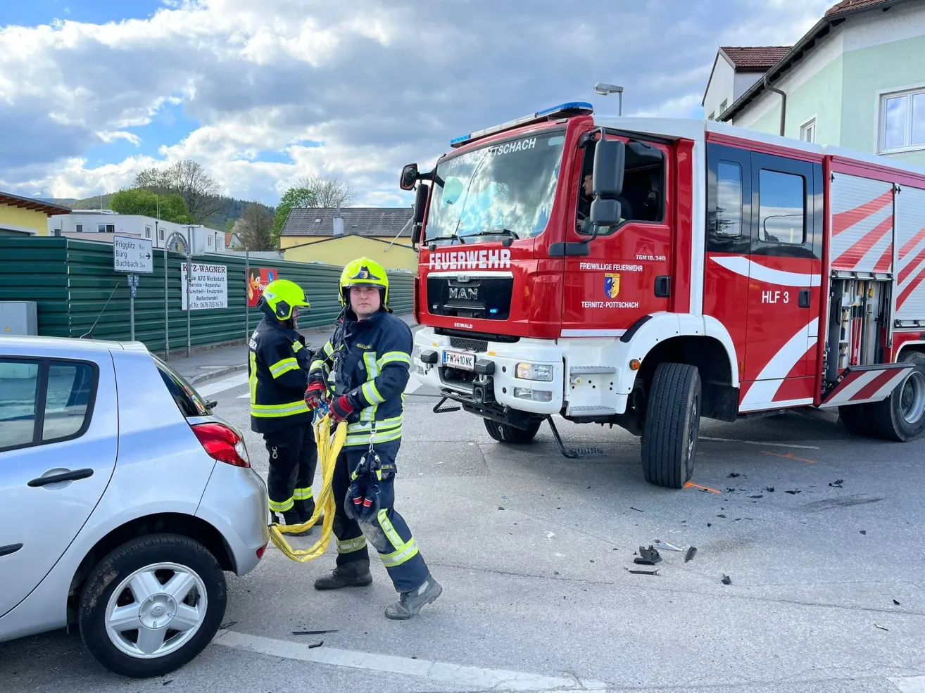 Two firefighters in yellow helmets assist a damaged car while a red fire truck is parked behind them on the street.