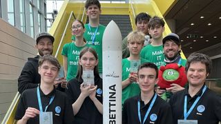 A group of young people wearing green shirts with Make It Matter on them are posing with a rocket labeled Bettina. Some hold awards, while others smile.