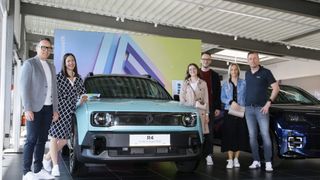 Several people stand next to a blue Renault electric car in a showroom. The car has a white plate reading R4.
