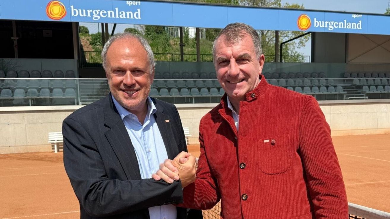 Two men stand smiling on a tennis court. One wears a black suit, the other a red corduroy jacket. Behind them is a blue sign with white text.