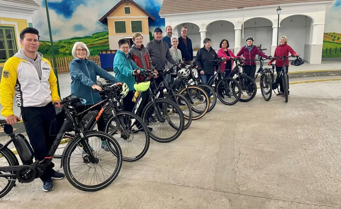 A group of people stands outside near bicycles, wearing helmets. Behind them, a building with an arch entrance and a mural is visible. They are smiling and posing for a photo.