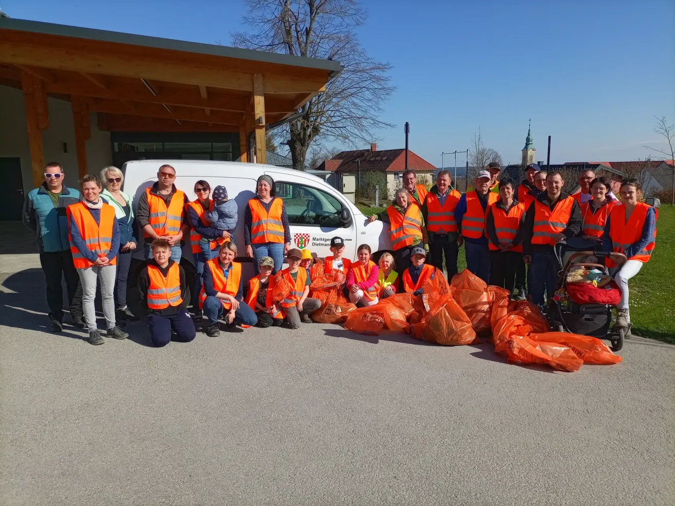 A group of people in reflective vests poses for a photo in front of a white van with a pile of orange bags. They are standing on the side of the road.