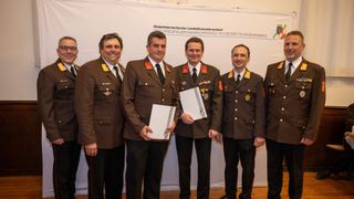 Five men in military uniforms stand next to each other and hold papers, smiling for a photo in front of a white backdrop.