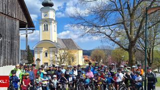 A group of people in cycling gear pose for a photo in front of a church with a clock tower, under a cloudy sky.
