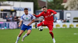 Two soccer players compete on a green field. One in red is kicking the ball, while the other in blue defends. The background features a blurred audience and a wall with advertisements.
