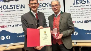 Two men in traditional German attire hold a certificate and a medal, smiling for a photo. The backdrop shows a poster with cow images and text in German.