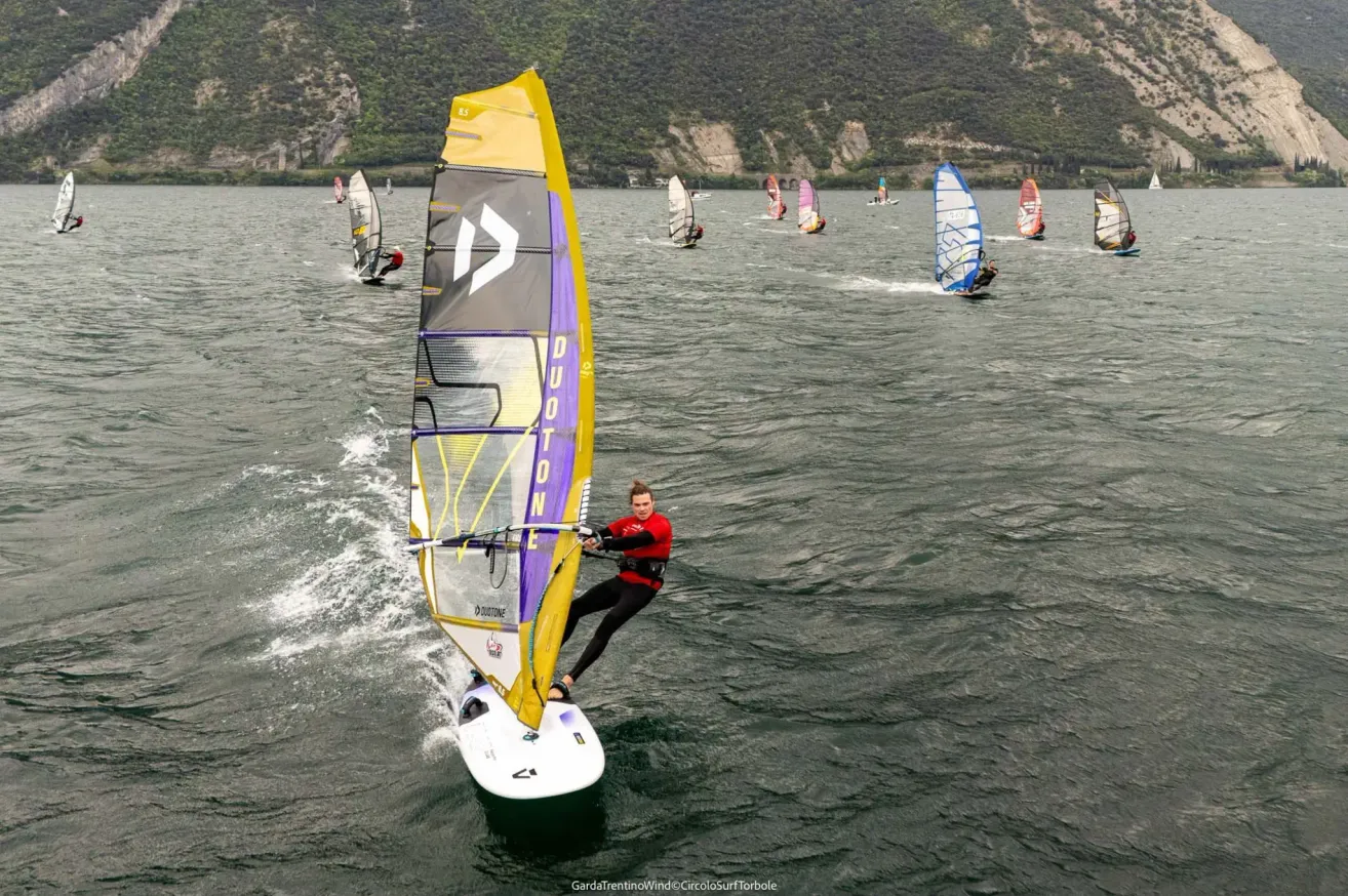 A group of windsurfers compete in a race on a lake with a mountainous backdrop. The closest surfer leans forward on a yellow and purple sailboard.