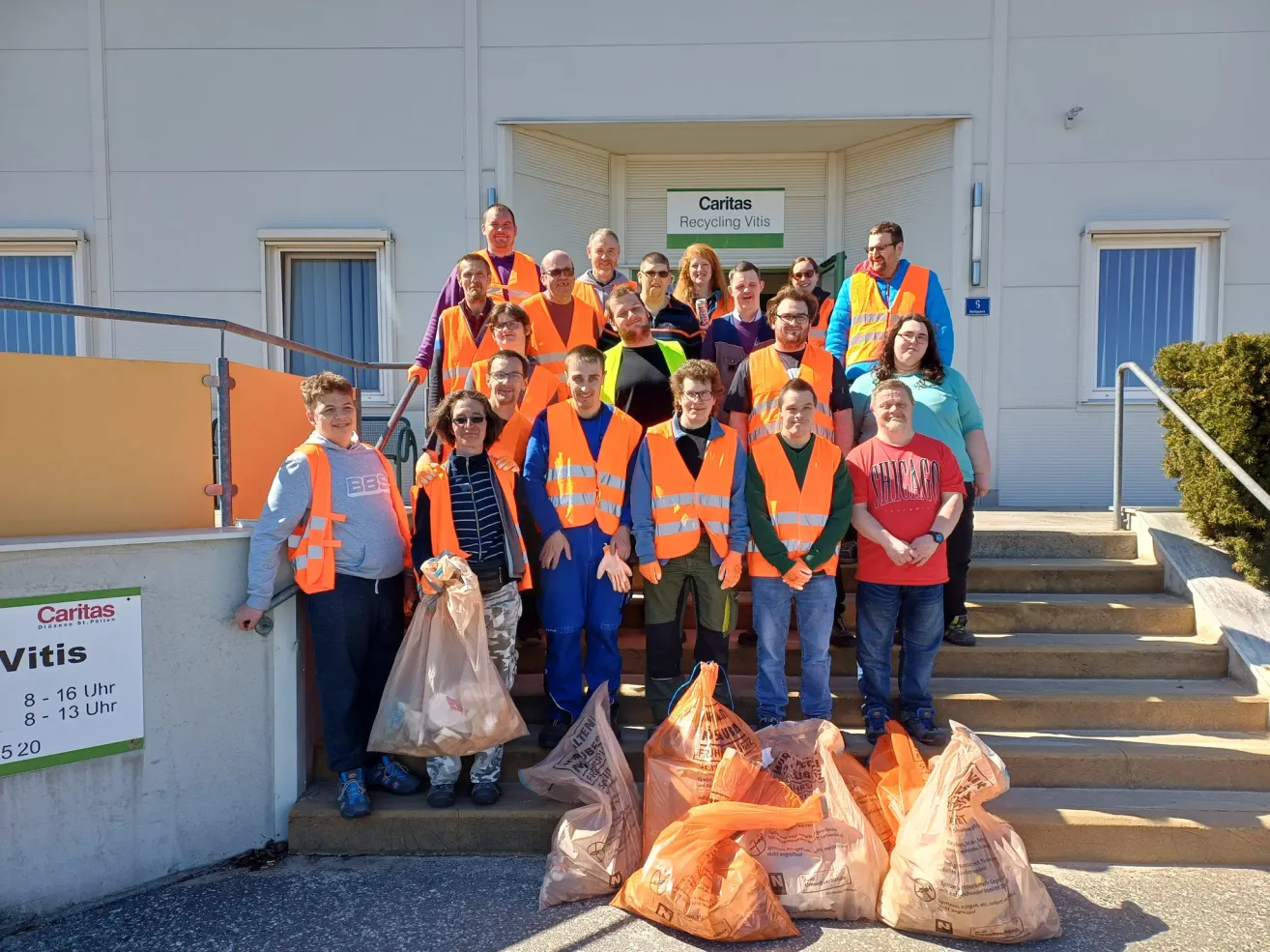 A group of people in orange vests standing on stairs, holding bags, with a sign for Caritas Recycling Vitis above them.