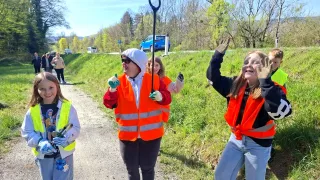 Four women in orange vests walk along a path, one holding a pitchfork. They are all smiling and appear to be enjoying themselves. In the background, there is a blue car and a road with a sign.