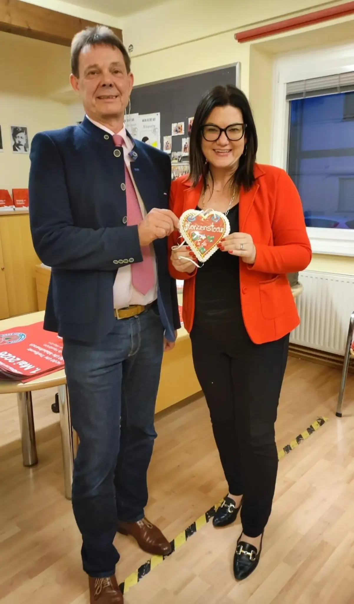 A man and a woman stand next to each other in a room. They hold a heart-shaped cookie labeled 'Herzenstoar'. Behind them is a table with red materials. A radiator is visible on the right.