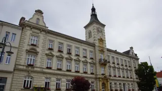 A large building with a clock tower stands tall against a cloudy sky. The building features many windows, each adorned with red flowers, and a yellow entrance.