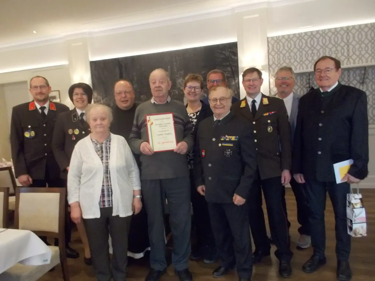 Group of military and civilians stand together. A man in the middle holds a certificate. They are dressed in formal attire.