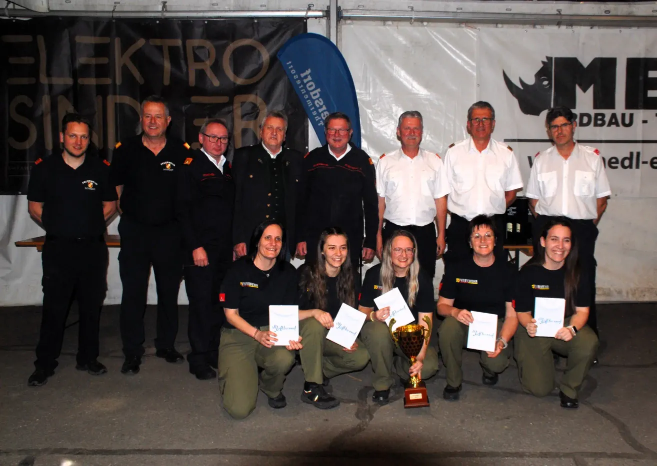 A group of people in uniforms, some holding awards, pose for a photo with a trophy in the center. A blue banner and a white banner are behind them.
