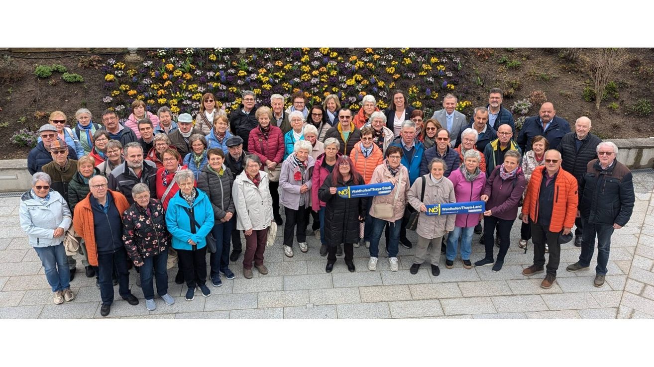 A large group of older people pose for a photo with blue signs. They stand in front of a flower wall.