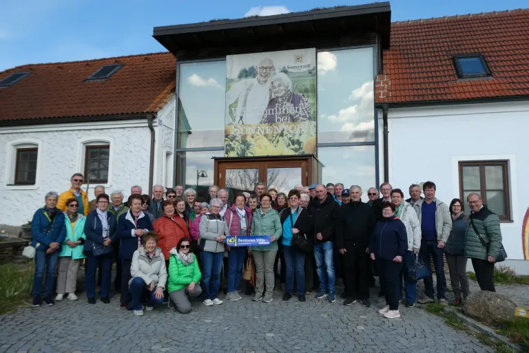 A group of people are posing for a photo outside a building with a large poster featuring elderly individuals.